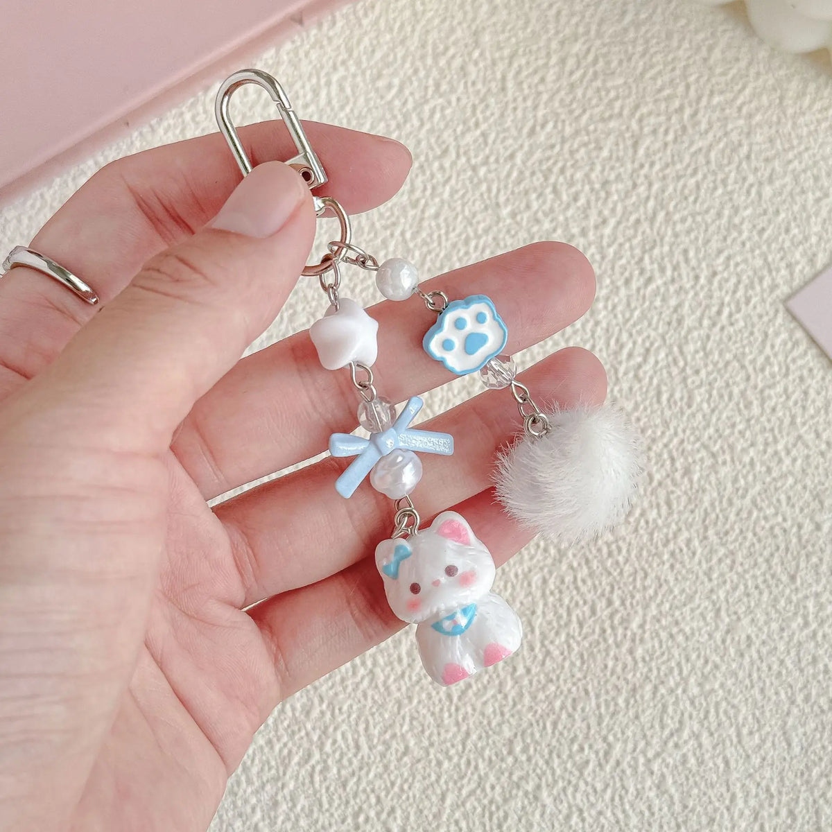 Blue cat bead strap charms held in a hand: paw bead, blue bow, pom-pom and cat